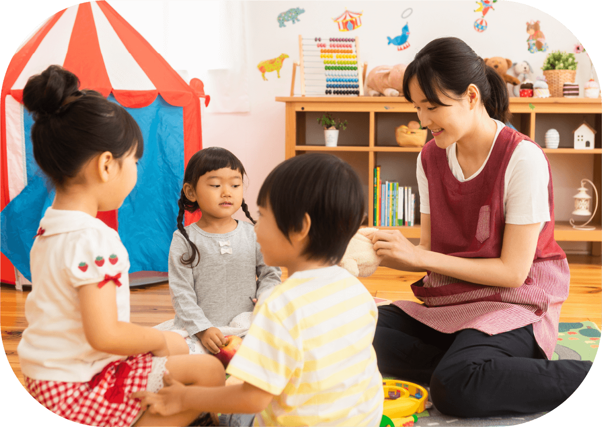 保育室で子供たちと向き合って座る保育士と遊び中の園児たち